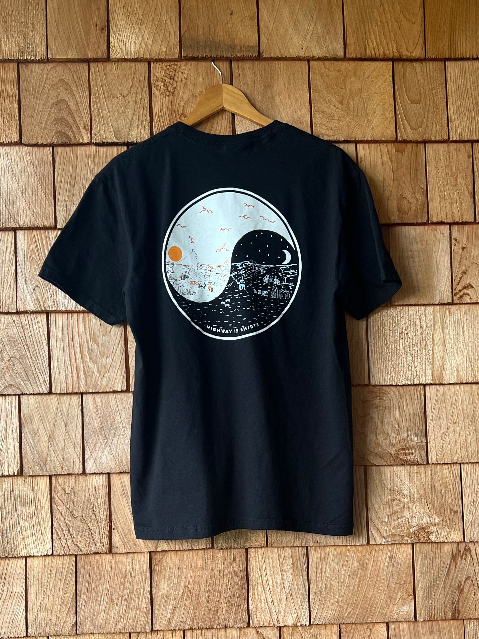 Black t-shirt with a white and gray design on a cedar shake wall.