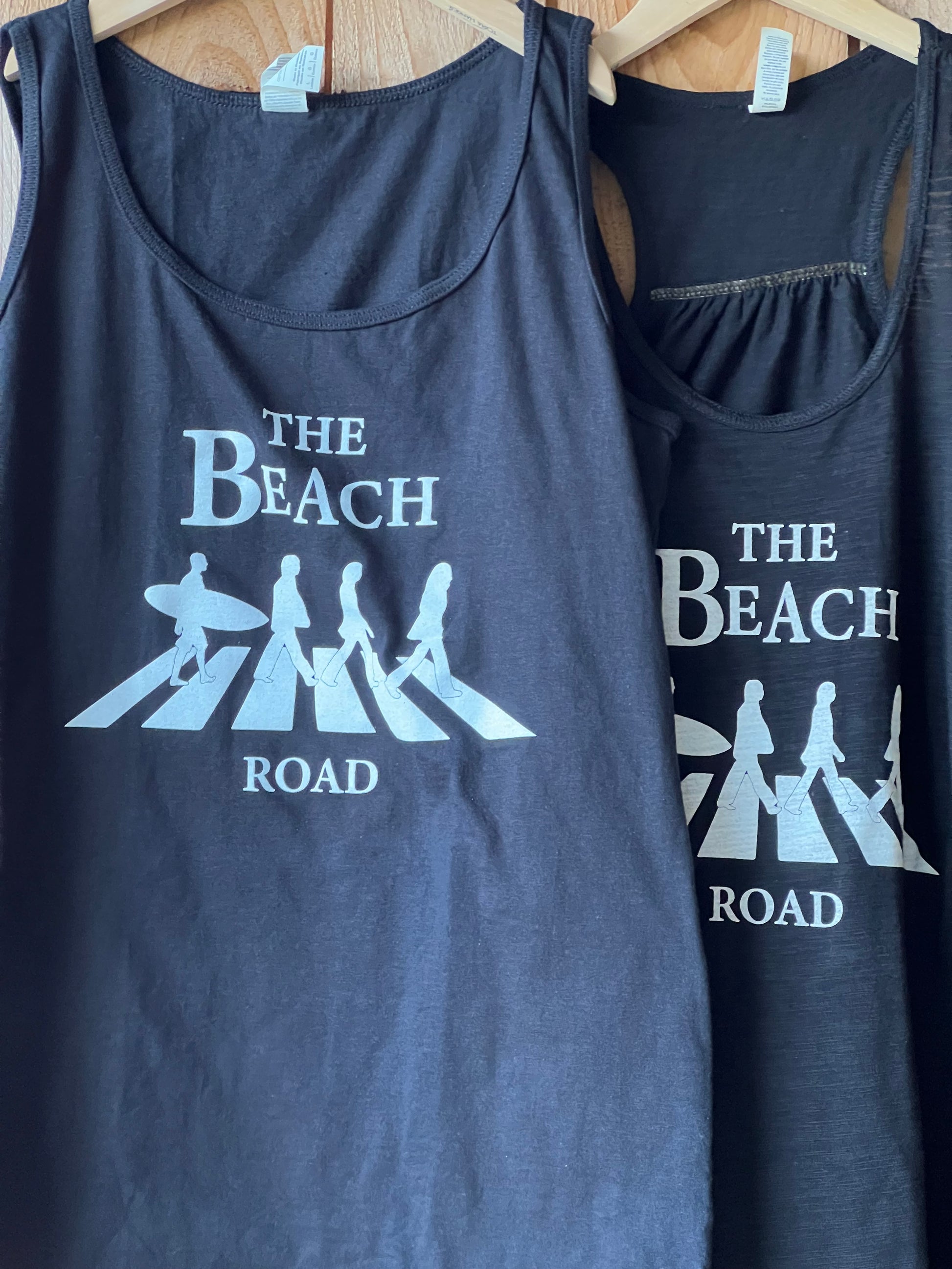 Two black blue tank tops with 'The Beach Road' logo on a wooden surface.
