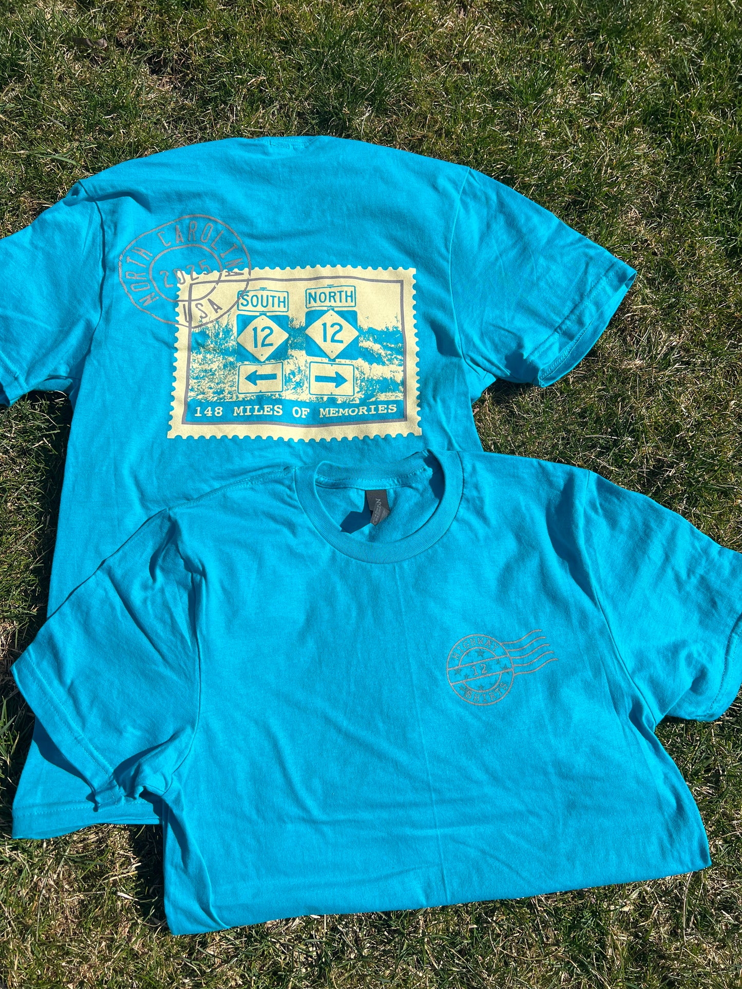 A blue colored t-shirt laid out on a grassy surface with a graphic design in the center that appears to be a postcard motif.