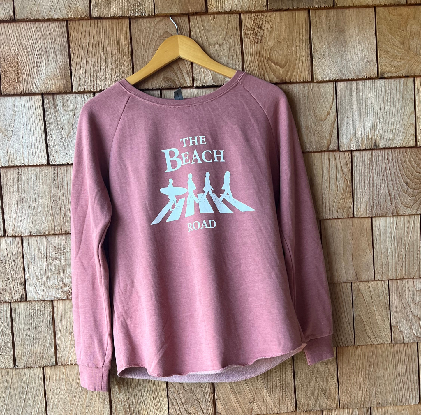 Pink sweatshirt with 'The Beach Road' design on a wooden wall